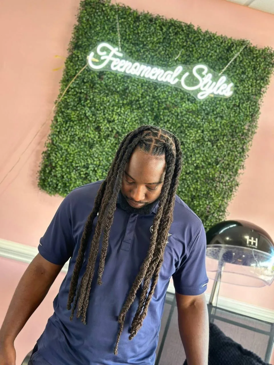 Man with long locs standing in front of the hedge wall and neon sign